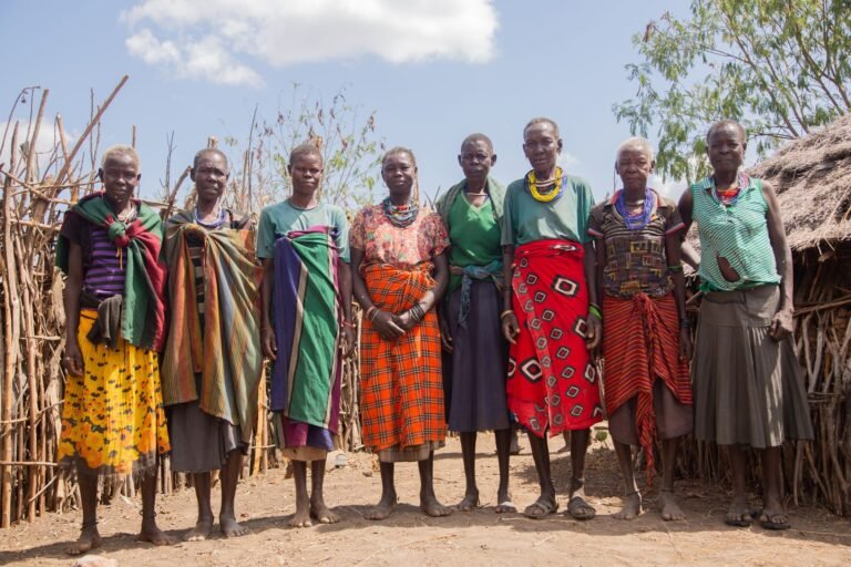 A group of women from an African tribe in traditional attire posing outdoors in their village.