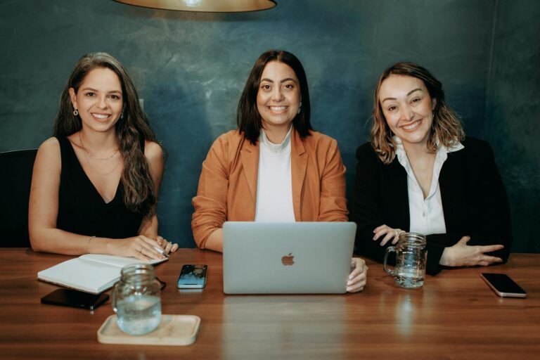 Three professional women sitting at a wooden desk in an office with laptops and notebooks.