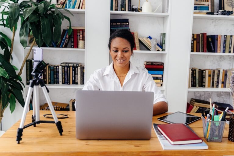 A young woman smiling while working on a laptop in a home office with bookshelves.