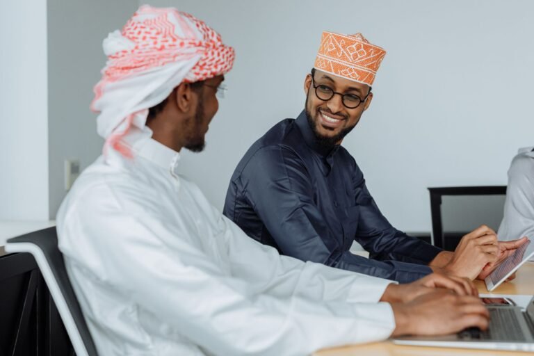 Men in traditional attire collaborating on laptops in an office, smiling and engaging.