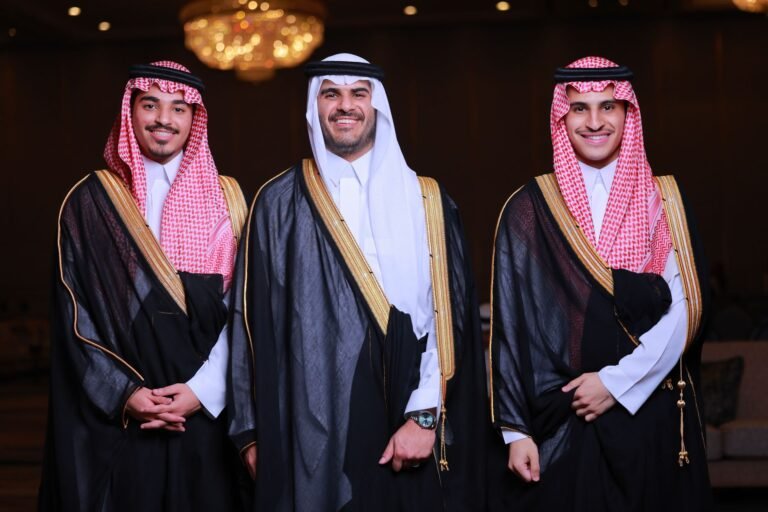 Three men in traditional Arabic robes and keffiyeh smiling at a formal indoor event.