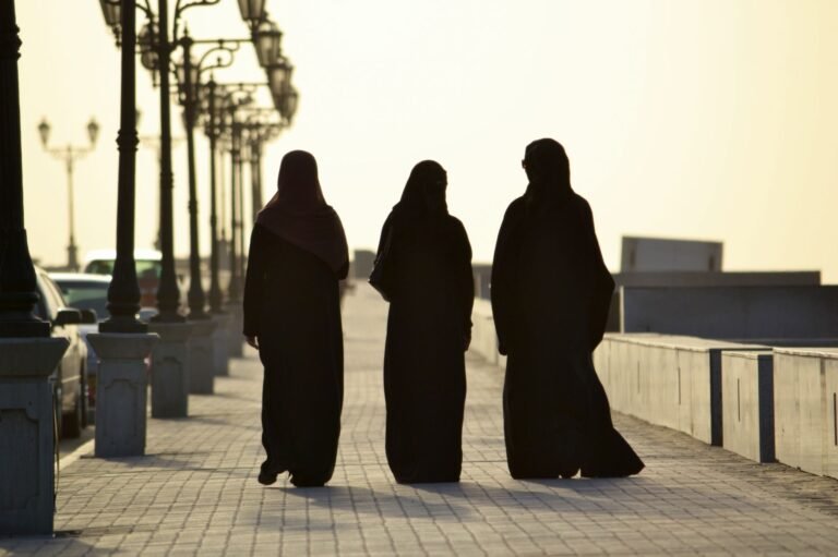 Silhouetted women in traditional clothing walking along a Muscat promenade at sunset.