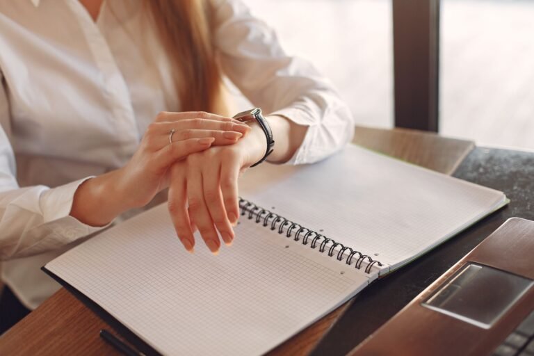 Unrecognizable crop female entrepreneur in formal clothes sitting at wooden table near laptop with copybook and checking time on wristwatch
